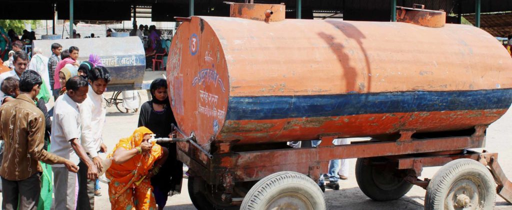 धूप में खड़े टैंकर से पानी पीते लोग। 